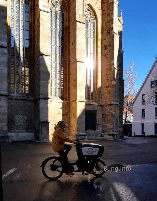 Radlerin vor hohen Kirchenfenstern, blauer Himmel, Asphalt nass, Schlagschatten