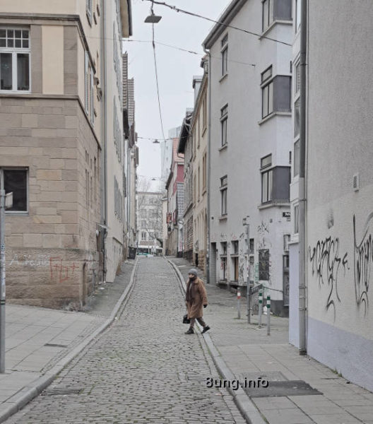 graue Häuser, graues Kopfsteinpflaster, grauer Himmel, Frau überquert die Strasse