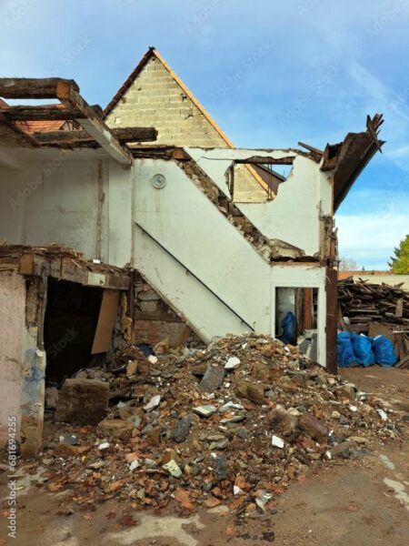 Ruine eines Fachwerkhauses, das abgerissen wurde. Eine Mauer steht noch, davor ein Steinhaufen mit Bauschutt, daneben blaue Müllsäcke und gestapelte Abbruchhölzer. Zerfetzte Balken ragen aus der Mauer.